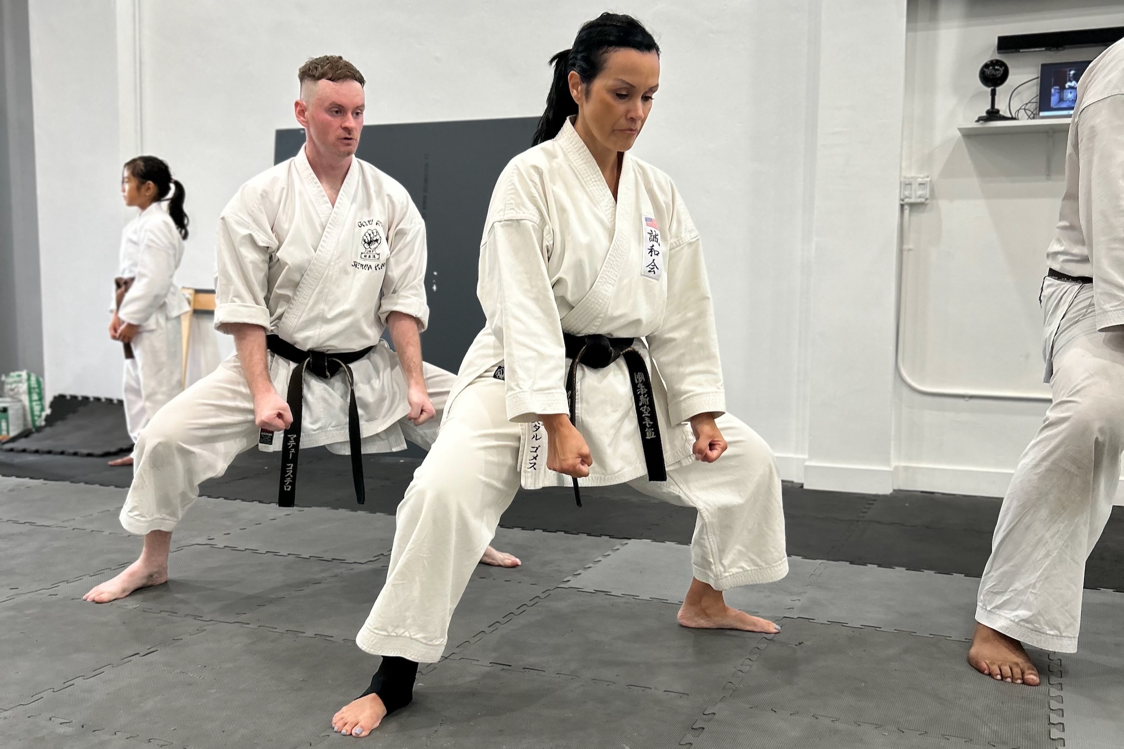 Adult martial arts students in white uniforms and black belts execute a low horse stance drill on grey training mats during an advanced class.