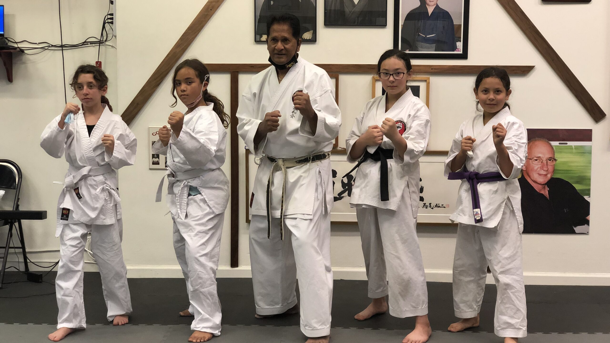A group photo of students and head instructor Vassie Shihan in traditional white karate gi, including youth students with purple and green belts, standing in front of a traditional dojo wall with framed historic martial arts portraits during our kids karate classes.
