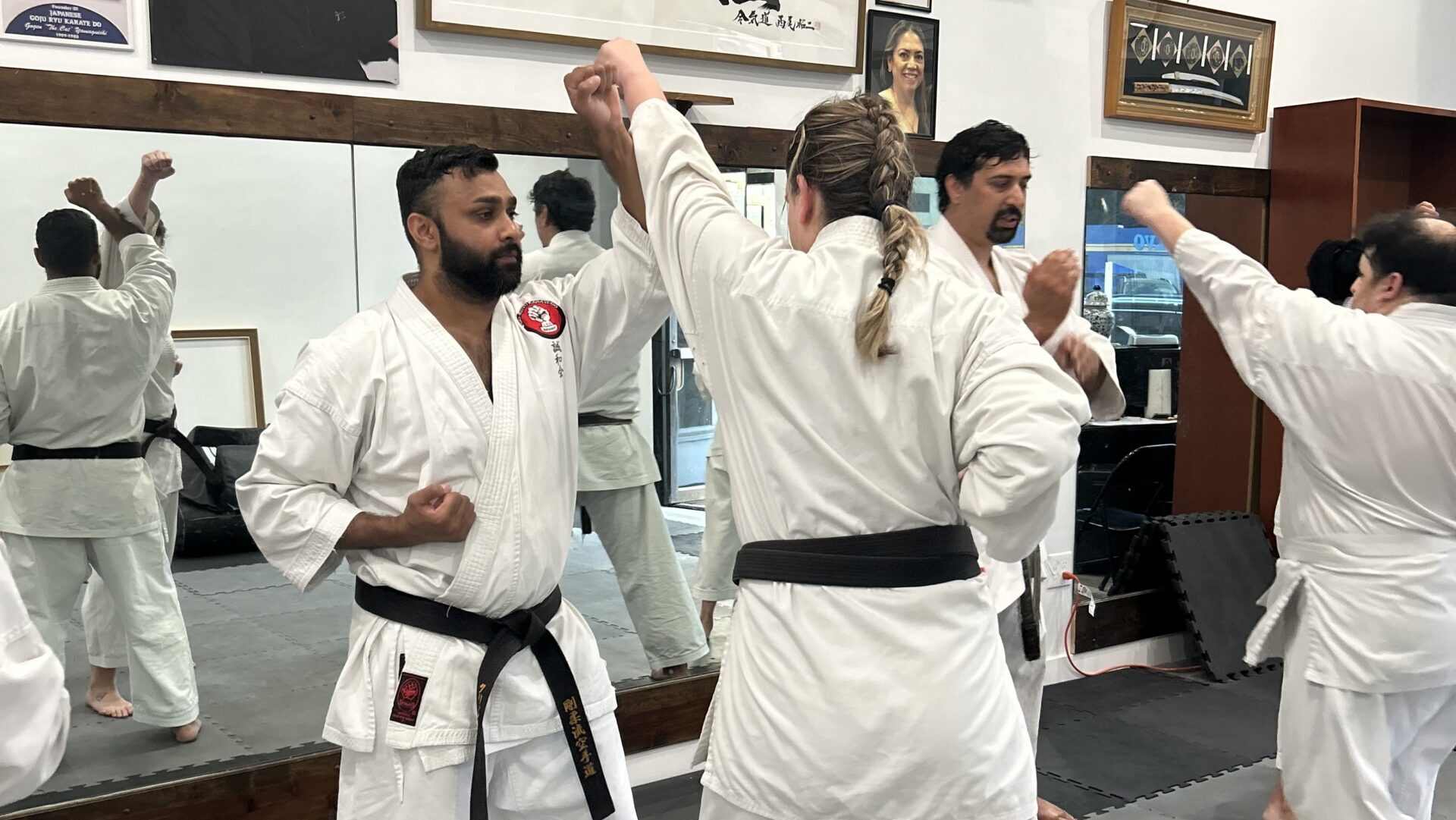 Two karate black belts practicing dynamic kumite sparring drills inside a Goju-Ryu dojo with instructors training in the background.