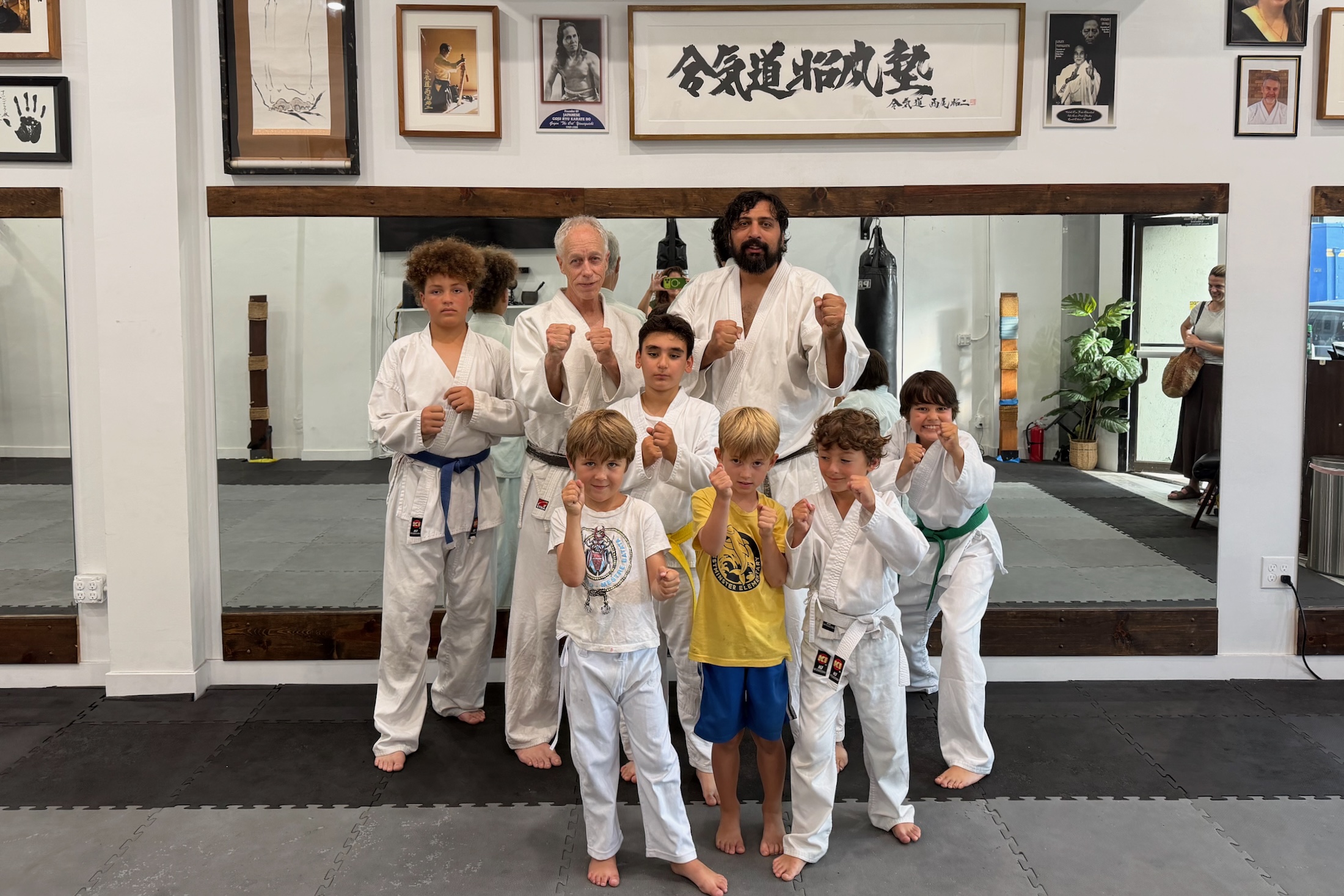 A group of smiling kids and instructors in traditional white karate uniforms posing with fists raised in front of a dojo wall with martial arts calligraphy.