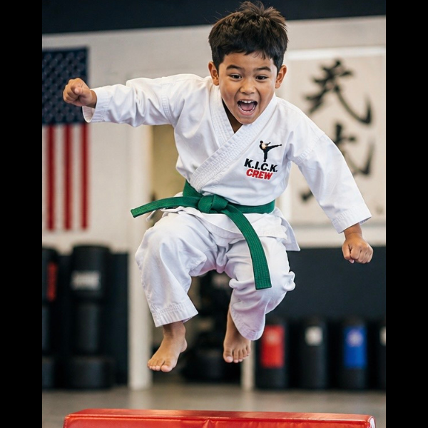 A young boy in a white karate gi jumping high in the air over a red pad with an excited smile