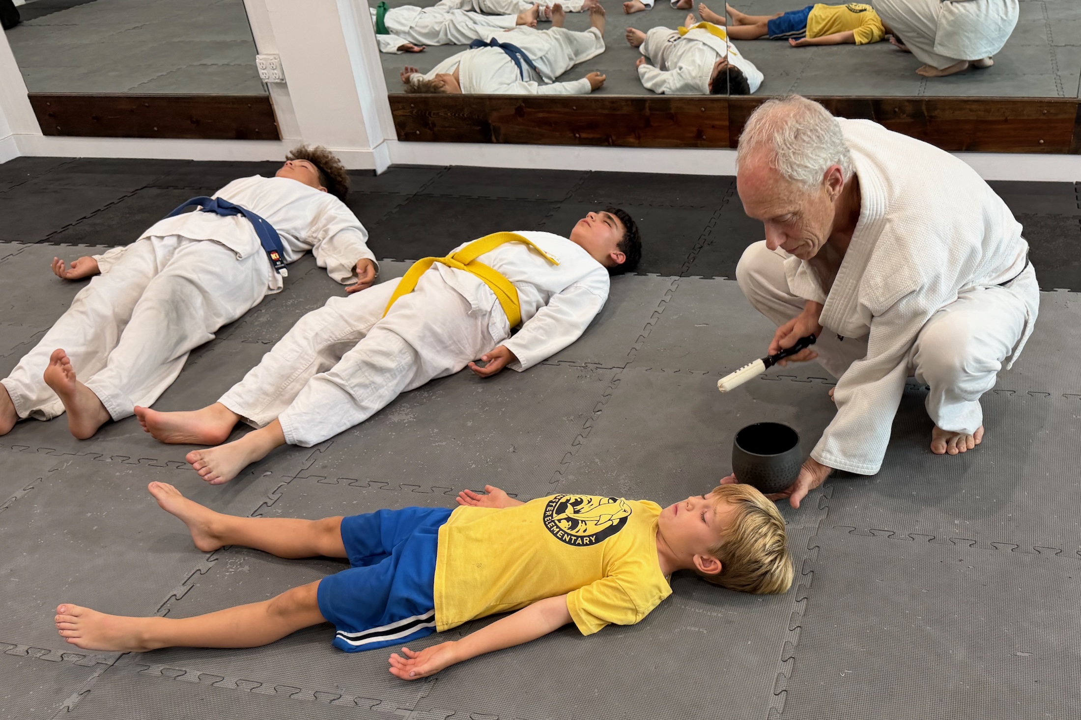 A karate instructor kneels on the mat with a traditional singing bowl next to children lying down practicing mindfulness meditation.