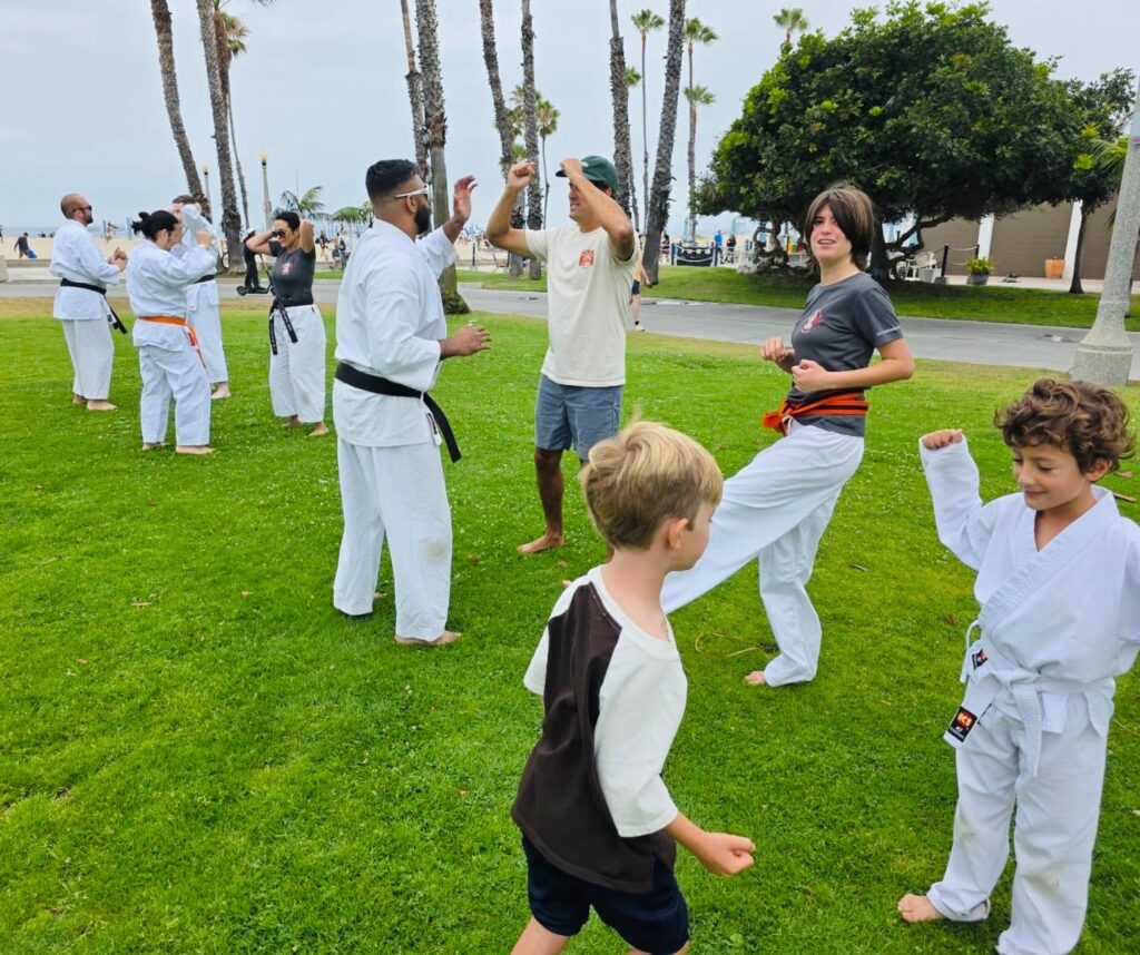 Outdoor Karate School Life Family Class Multi-generational karate school life showcasing adult and youth program students training together on the grass under palm trees.
