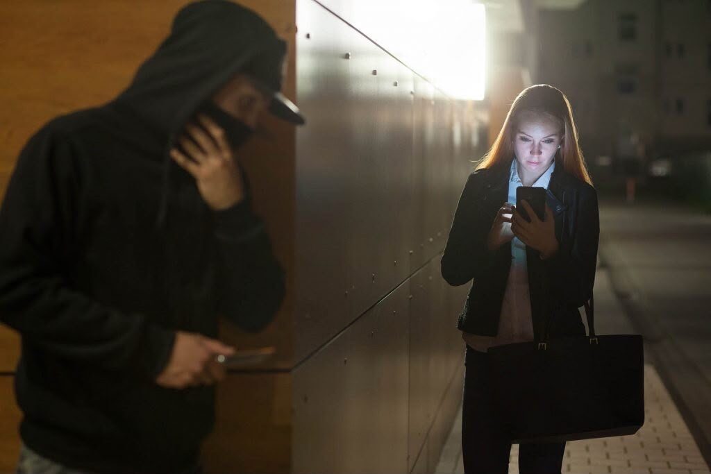 A distracted woman looks down at her glowing smartphone screen at night on a city street, unaware of a person in a dark hoodie watching her from the foreground shadow.