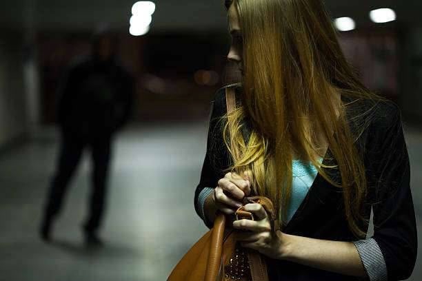 A woman clutching her handbag looks back over her shoulder into a dark, blurry underground tunnel where a shadowed figure walks toward her.