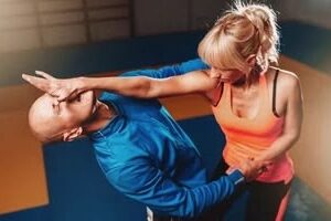 A woman in an orange tank top practices a self-defense palm strike to a man's nose and face on blue and orange training mats.